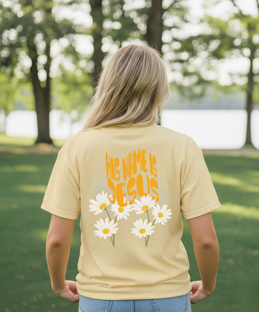 Person wearing a yellow t-shirt with 'His Name is Jesus' and daisy flowers outdoors.